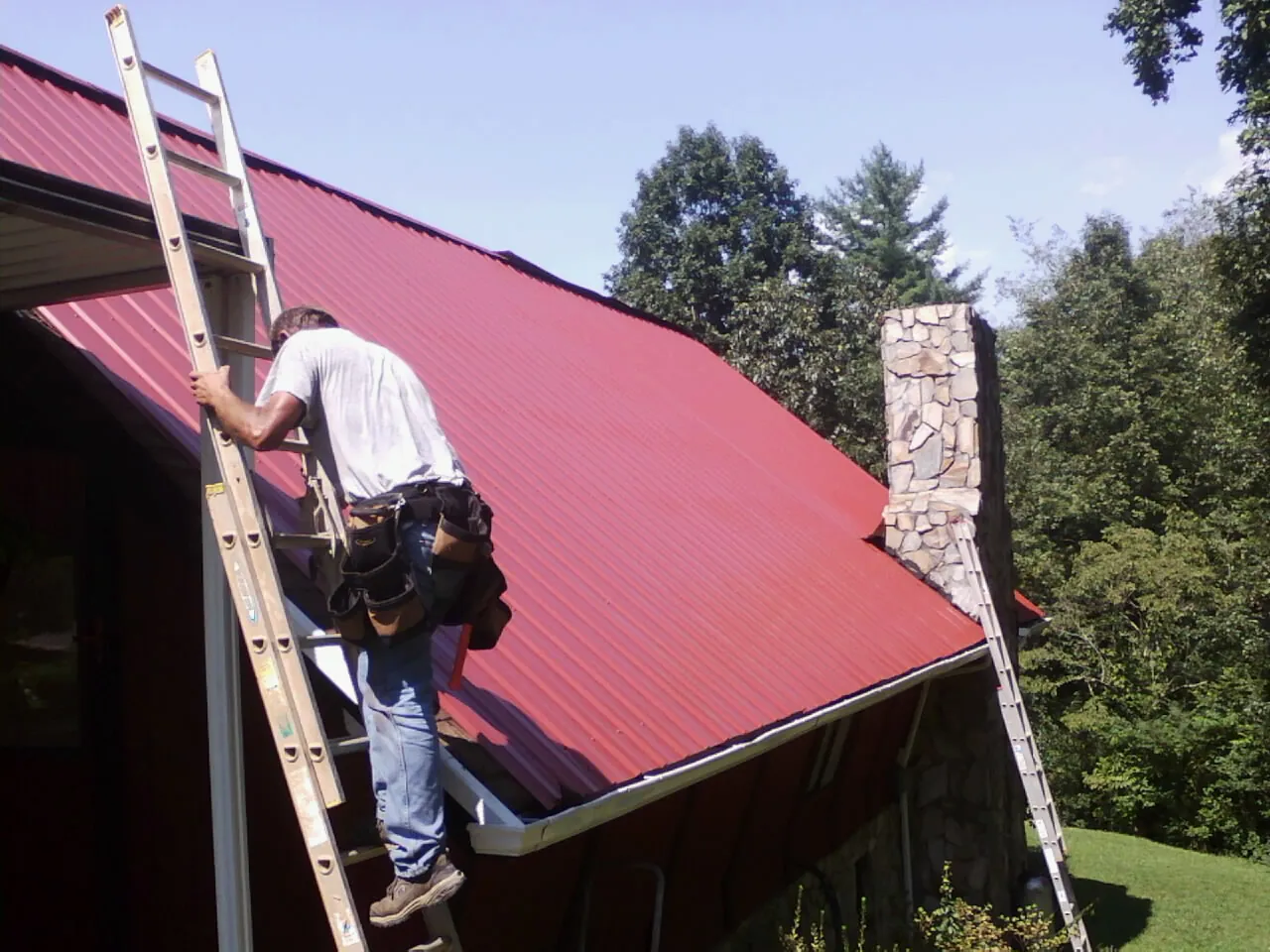 Roofer on a ladder beside a red metal roof and stone chimney — Soffit & Fascia Repair by Roofing Brothers in Eutaw